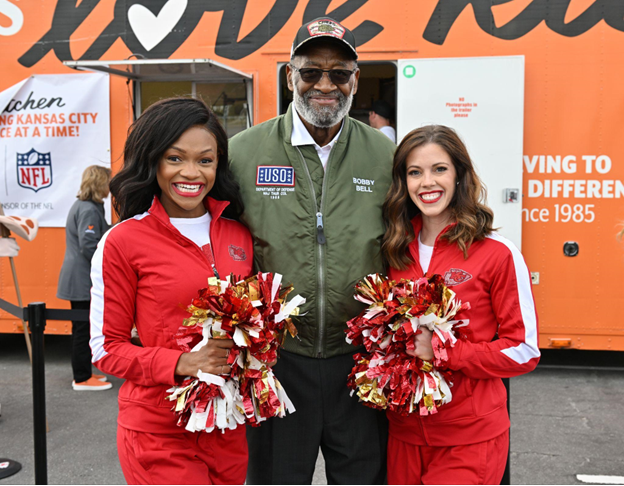 Kansas City Chiefs cheerleaders with pom poms with Hall of Fame NFL Player Bobby Bell wearing a green jacket and black hat smiling. 