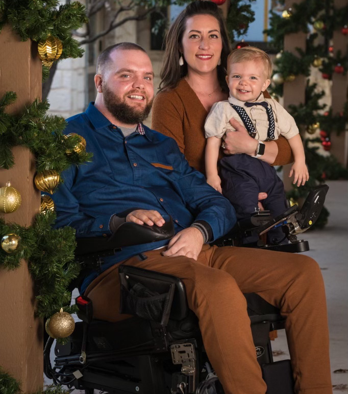 A man in a wheelchair and his wife and son pose in front of a winter holiday display.
