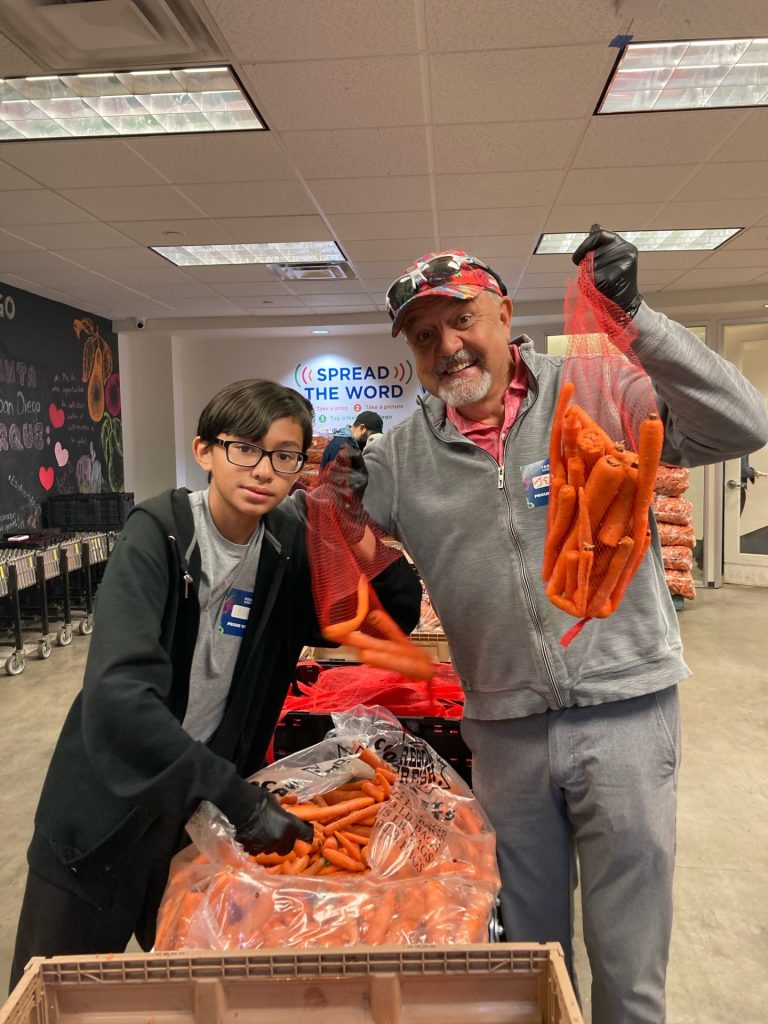 Boy with glasses poses with older man in baseball cap and gray sweatshirt holding bags of carrots during volunteer event.