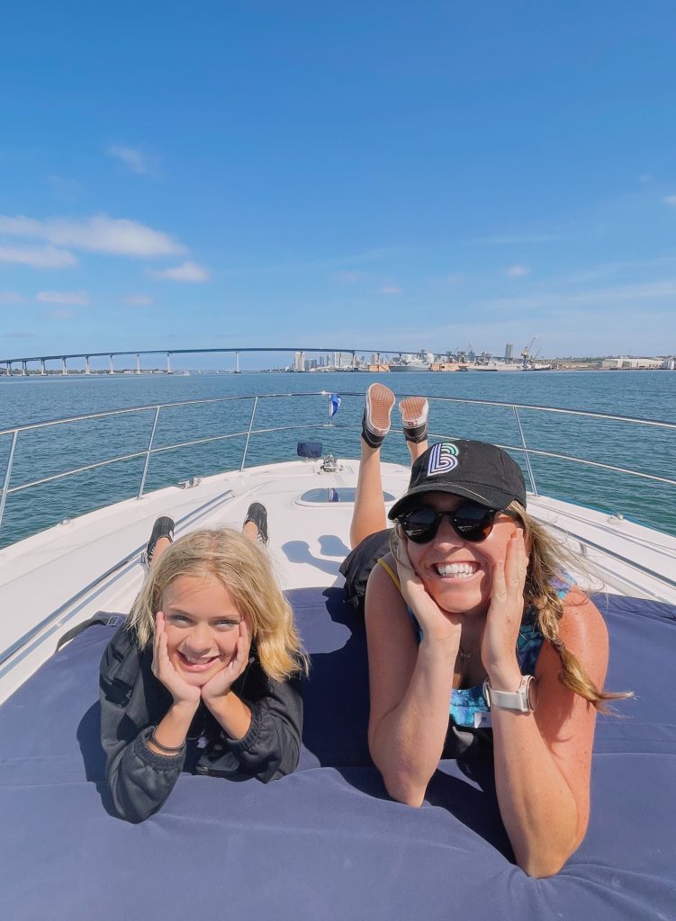 Girl and young woman in sunglasses and baseball cap pose on the front of a boat on the San Diego Bay near the Coranado Bridge.