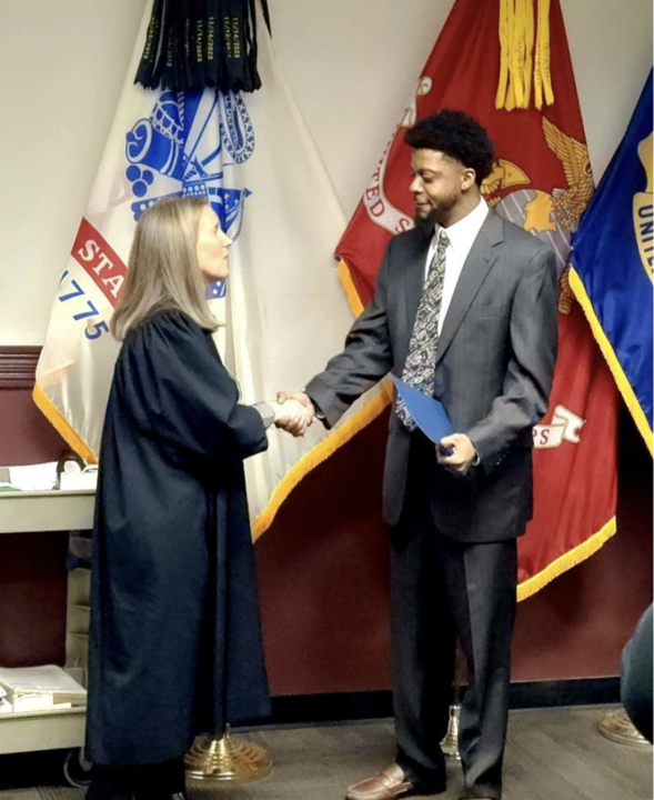 female judge in black robe shakes hands with man in gray suit and tie who completed the program standing in front of Maryland state flag.
